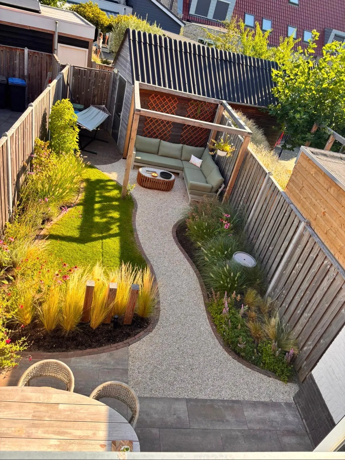 Dezelfde tuin na de aanleg, met een gebogen grindpad, gazon, beplanting en een loungehoek onder een pergola