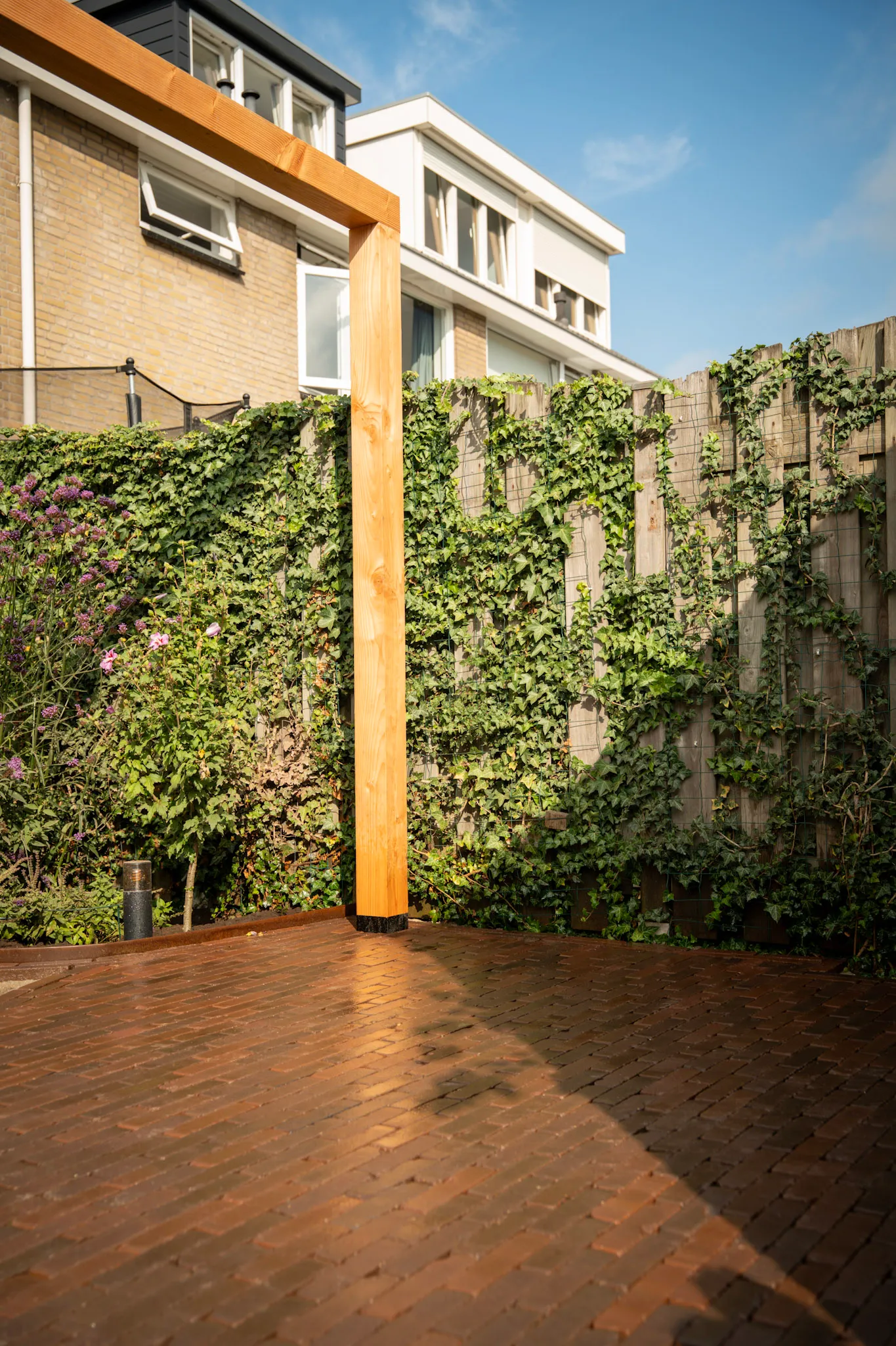 Houten pergola op een klinkerterras naast een groene schutting