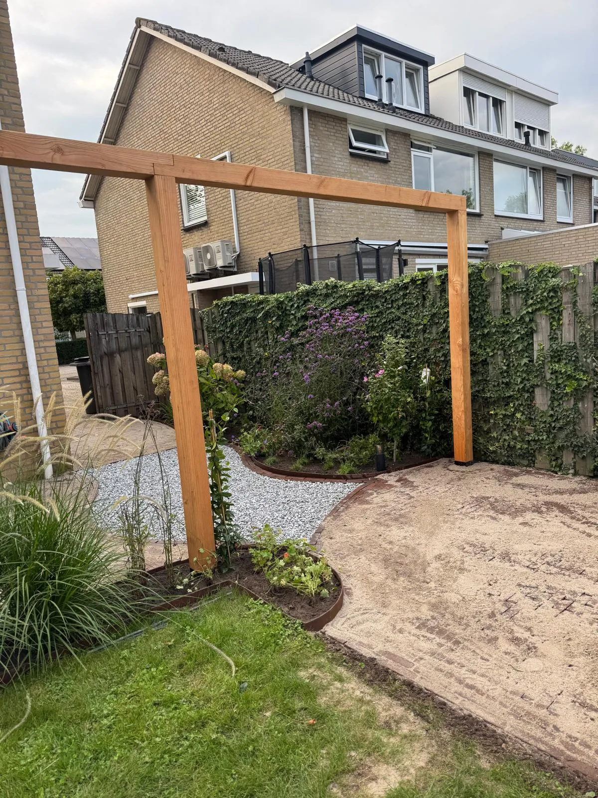 Houten pergola tussen bestaande bloemenborders en een gebogen grindpad