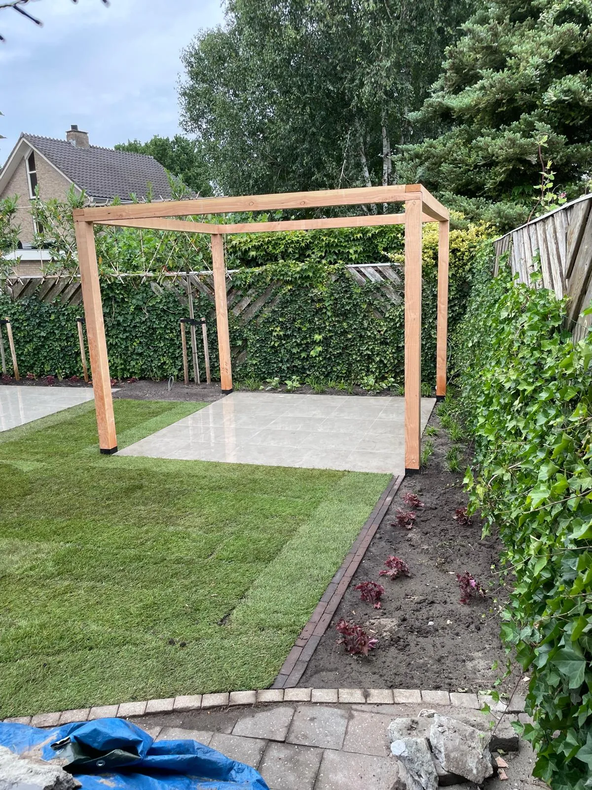 Houten pergola boven een betegelde zitplek, naast gazon en nieuwe borders